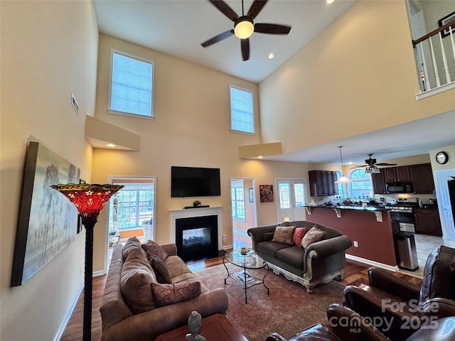 living room featuring a towering ceiling, wood finished floors, visible vents, a ceiling fan, and a glass covered fireplace