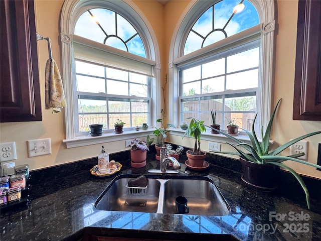 kitchen with dark brown cabinetry, a sink, dark stone counters, and a healthy amount of sunlight