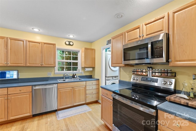 kitchen featuring dark countertops, stacked washing maching and dryer, a sink, light wood finished floors, and appliances with stainless steel finishes