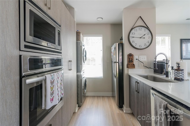 kitchen with light wood-style flooring, a sink, modern cabinets, stacked washing maching and dryer, and appliances with stainless steel finishes