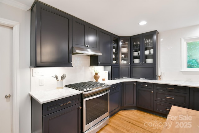 kitchen featuring glass insert cabinets, tasteful backsplash, under cabinet range hood, stainless steel gas range oven, and light wood finished floors