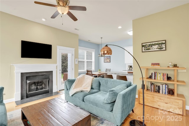 living room with ceiling fan with notable chandelier, a fireplace with raised hearth, recessed lighting, baseboards, and hardwood / wood-style flooring