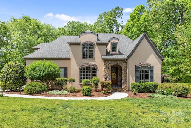 french country home featuring a front lawn, stone siding, stucco siding, and roof with shingles
