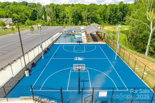 view of sport court featuring community basketball court, fence, a wooded view, and playground community