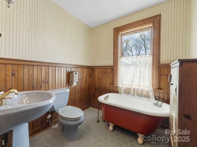 bathroom with a freestanding bath, a sink, toilet, wainscoting, and wallpapered walls