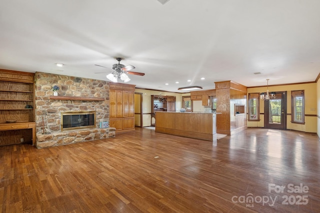unfurnished living room featuring dark wood-style flooring, a stone fireplace, recessed lighting, crown molding, and ceiling fan with notable chandelier