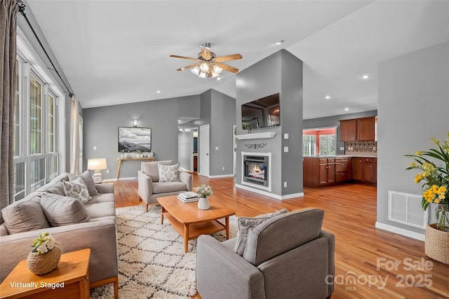 living area featuring ceiling fan, light wood-type flooring, a glass covered fireplace, visible vents, and lofted ceiling
