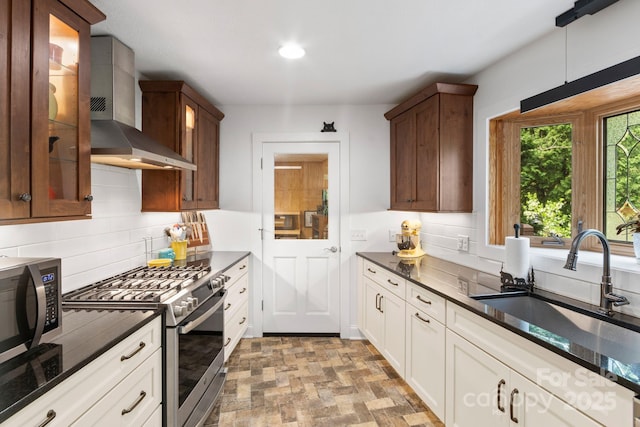 kitchen featuring wall chimney range hood, a sink, stainless steel gas range oven, decorative backsplash, and glass insert cabinets