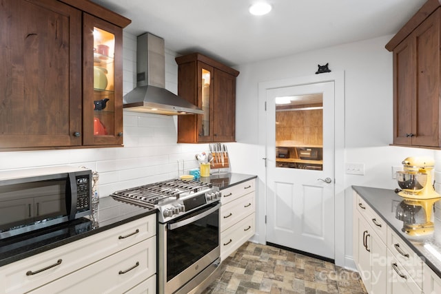 kitchen with appliances with stainless steel finishes, wall chimney exhaust hood, dark stone counters, backsplash, and glass insert cabinets