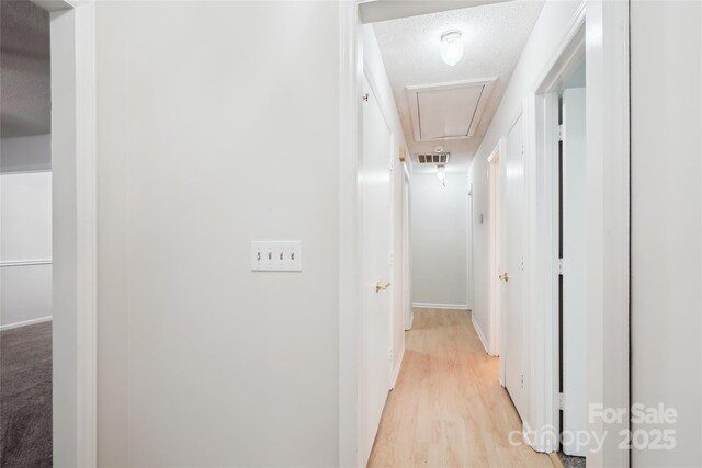 hall with attic access, a textured ceiling, visible vents, baseboards, and light wood-style floors