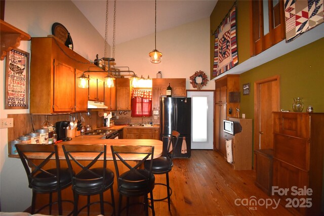 kitchen featuring brown cabinets, high vaulted ceiling, freestanding refrigerator, dark wood-type flooring, and under cabinet range hood