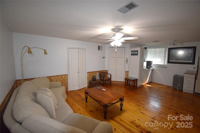 living area featuring visible vents, light wood finished floors, ceiling fan, and a wainscoted wall