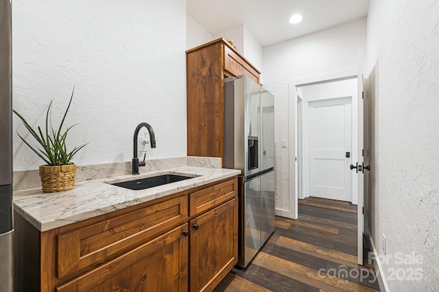 kitchen with stainless steel fridge, a sink, light stone countertops, a textured wall, and dark wood-style floors