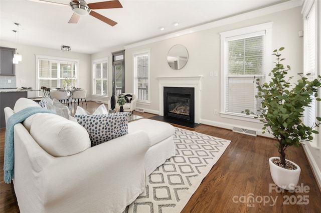 living area featuring a ceiling fan, dark wood-type flooring, a fireplace with flush hearth, baseboards, and ornamental molding
