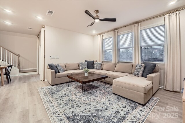 living area featuring ceiling fan, light wood-style floors, baseboards, visible vents, and stairway