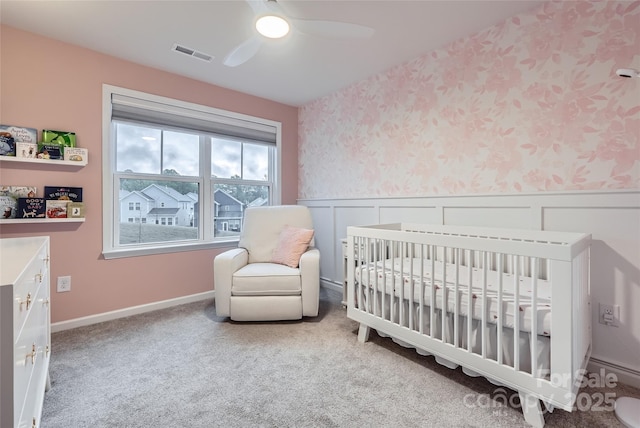 carpeted bedroom featuring a ceiling fan, visible vents, a nursery area, a wainscoted wall, and wallpapered walls