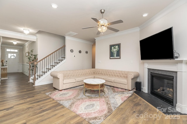 living room with crown molding, a ceiling fan, a glass covered fireplace, stairway, and wood finished floors