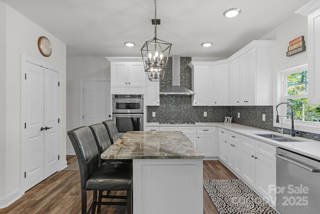 kitchen featuring appliances with stainless steel finishes, a sink, a chandelier, wall chimney exhaust hood, and white cabinetry