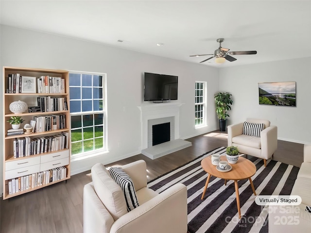 living area featuring a fireplace with raised hearth, baseboards, a ceiling fan, and wood finished floors