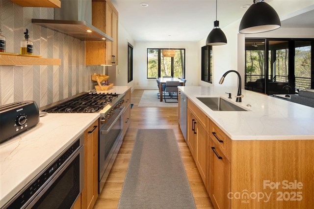 kitchen featuring light wood finished floors, a sink, brown cabinets, stainless steel appliances, and wall chimney exhaust hood