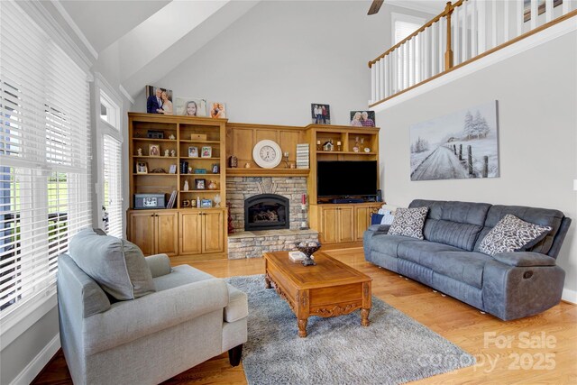 living area featuring high vaulted ceiling, a ceiling fan, a stone fireplace, and light wood-type flooring