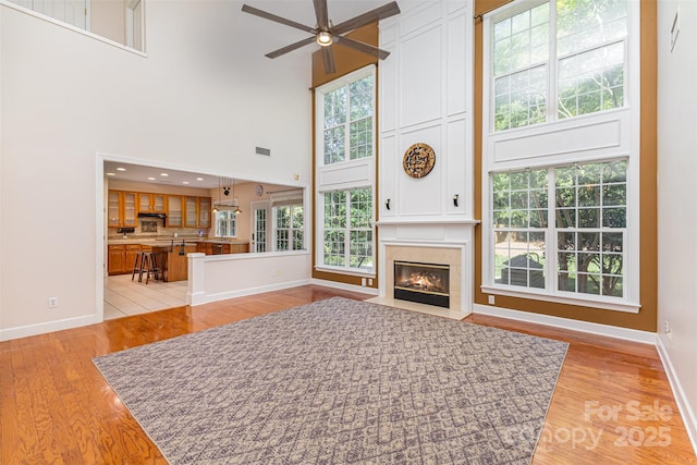 living area featuring a ceiling fan, baseboards, light wood-style flooring, a towering ceiling, and a high end fireplace