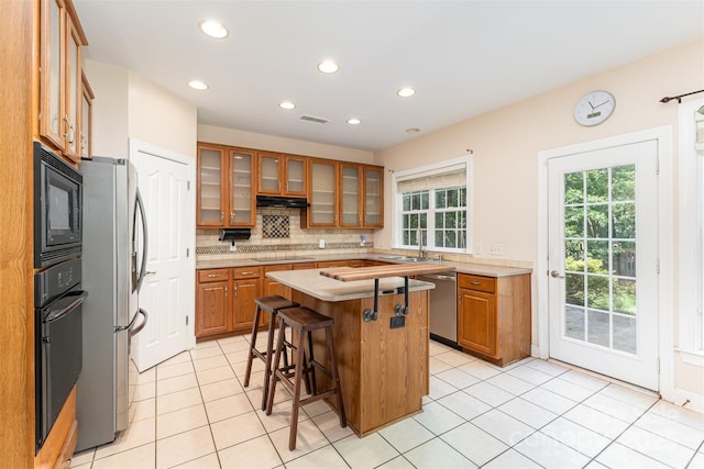 kitchen with black appliances, brown cabinetry, a kitchen bar, under cabinet range hood, and recessed lighting
