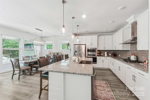 kitchen featuring wood finished floors, wall chimney exhaust hood, stainless steel appliances, and a kitchen island with sink