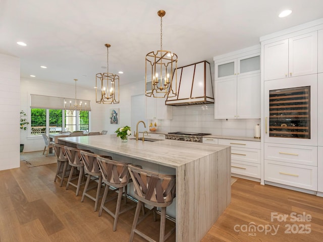 kitchen featuring a notable chandelier, a sink, a kitchen island with sink, beverage cooler, and custom range hood