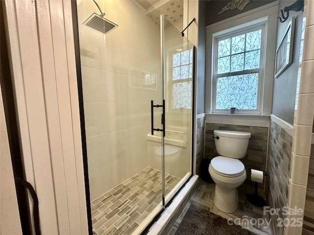 bathroom featuring toilet, a stall shower, a wainscoted wall, and tile walls
