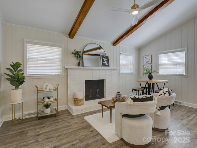 living area featuring wood finished floors, visible vents, a ceiling fan, lofted ceiling with beams, and a brick fireplace