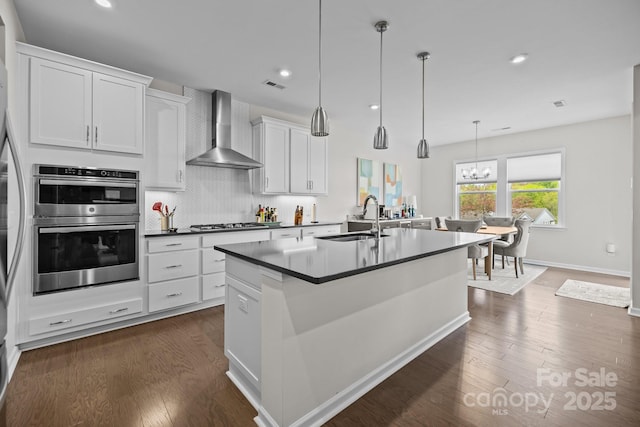 kitchen with a sink, a kitchen island with sink, wall chimney exhaust hood, appliances with stainless steel finishes, and visible vents