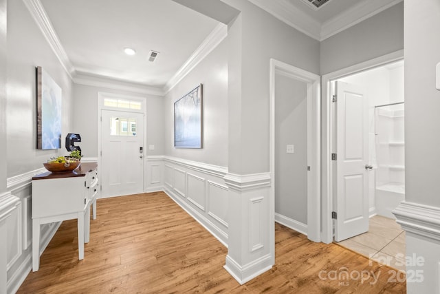 foyer with crown molding, light wood finished floors, a decorative wall, a wainscoted wall, and visible vents