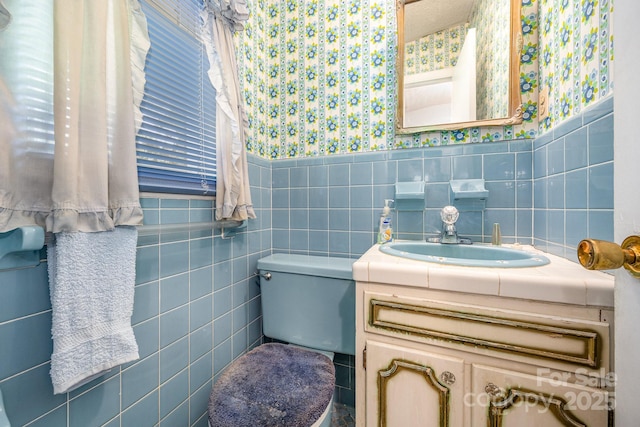half bath featuring wallpapered walls, a wainscoted wall, tile walls, toilet, and vanity