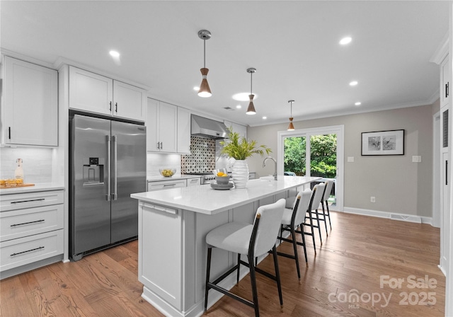 kitchen featuring high end refrigerator, wall chimney range hood, tasteful backsplash, ornamental molding, and an island with sink