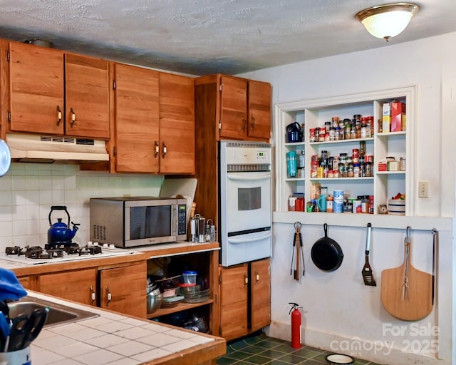 kitchen featuring tile countertops, brown cabinets, a warming drawer, stainless steel microwave, and under cabinet range hood