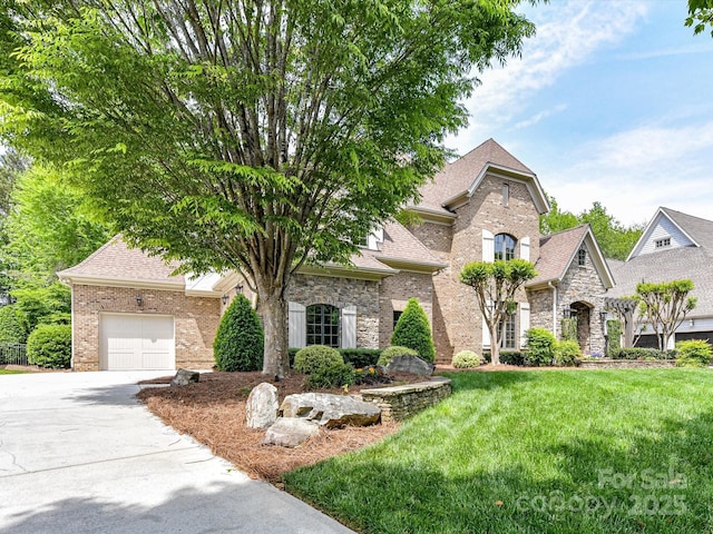 french country inspired facade with roof with shingles, driveway, brick siding, an attached garage, and a front yard