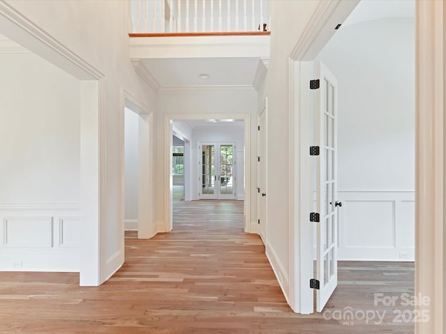 corridor featuring french doors, light wood-type flooring, wainscoting, a decorative wall, and ornamental molding