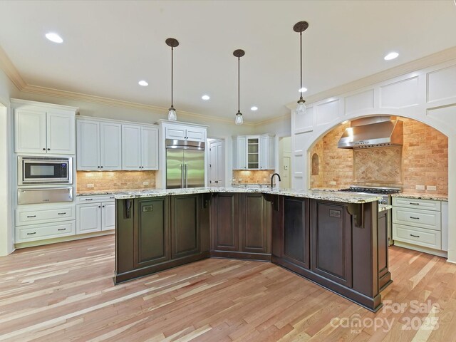 kitchen with wall chimney range hood, built in appliances, a warming drawer, backsplash, and white cabinetry