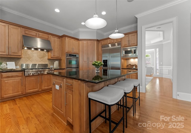 kitchen with wall chimney exhaust hood, built in appliances, a kitchen island, a breakfast bar area, and light wood-type flooring