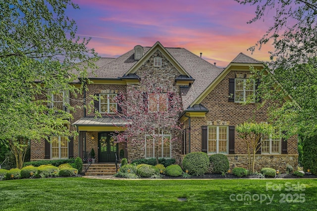 view of front of home with stone siding, roof with shingles, french doors, brick siding, and a yard