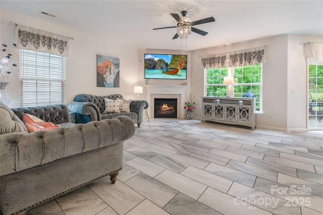 living room with baseboards, visible vents, a ceiling fan, and a warm lit fireplace