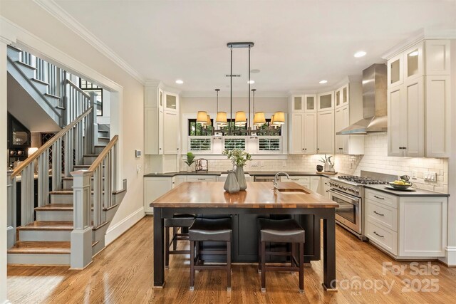 kitchen featuring wall chimney exhaust hood, a kitchen island with sink, white cabinets, high end range, and wooden counters
