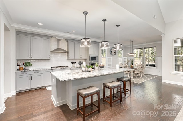kitchen with an island with sink, stainless steel appliances, a breakfast bar, gray cabinets, and wall chimney exhaust hood