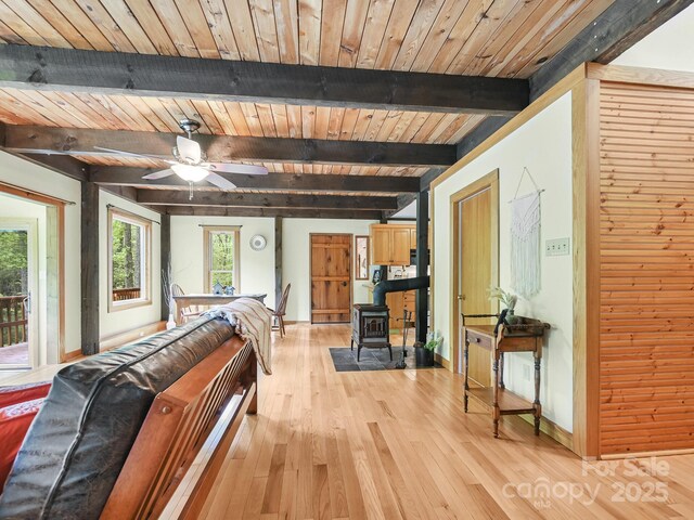 living area with ceiling fan, a wood stove, baseboards, beamed ceiling, and light wood-style floors