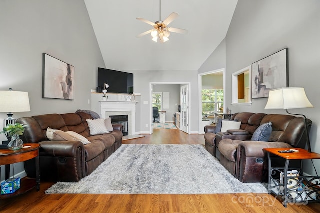 living area with a glass covered fireplace, wood finished floors, baseboards, high vaulted ceiling, and a ceiling fan
