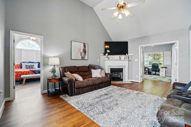 living area with a fireplace with raised hearth, high vaulted ceiling, a ceiling fan, baseboards, and wood finished floors