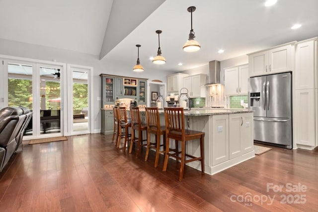 kitchen featuring wall chimney range hood, a breakfast bar, glass insert cabinets, dark wood finished floors, and stainless steel refrigerator with ice dispenser