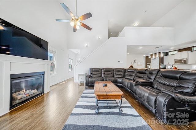 living room featuring a towering ceiling, wood finished floors, a ceiling fan, and a glass covered fireplace