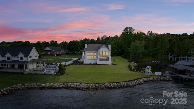 back of property at dusk featuring a lawn, a water view, an outdoor pool, and a chimney